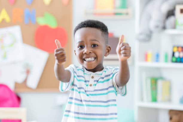 An adorable preschool age little boy smiles for the camera as he stands in his preschool classroom and gives a thumbs up. He loves school!